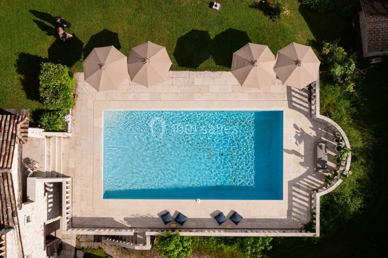 Vue aérienne d'une piscine rectangulaire entourée de parasols beige et de chaises longues sur une terrasse en pierre.