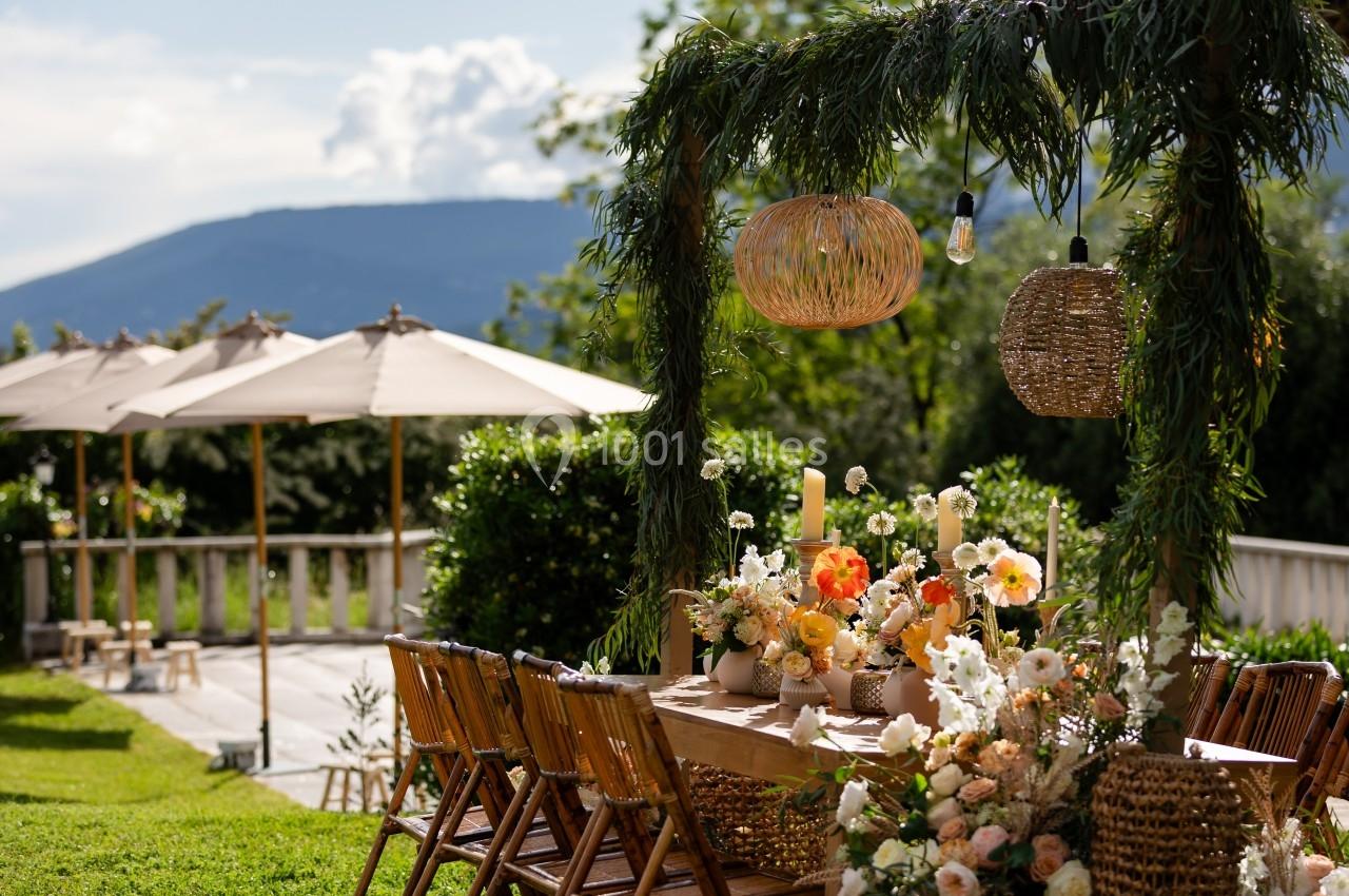 Table décorée avec des fleurs et des bougies sous une pergola ornée de feuillage, avec vue sur un jardin ensoleillé.
