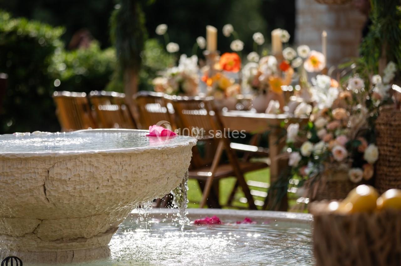 Fontaine en pierre avec de l'eau qui coule, entourée de fleurs et de tables décorées en extérieur.
