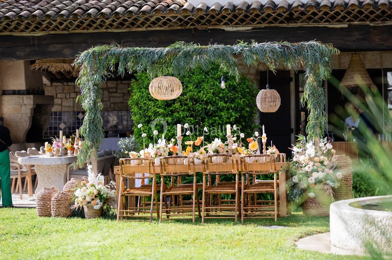 Table en bois décorée de fleurs et bougies sous une pergola ornée de feuillage, dans un jardin ensoleillé.