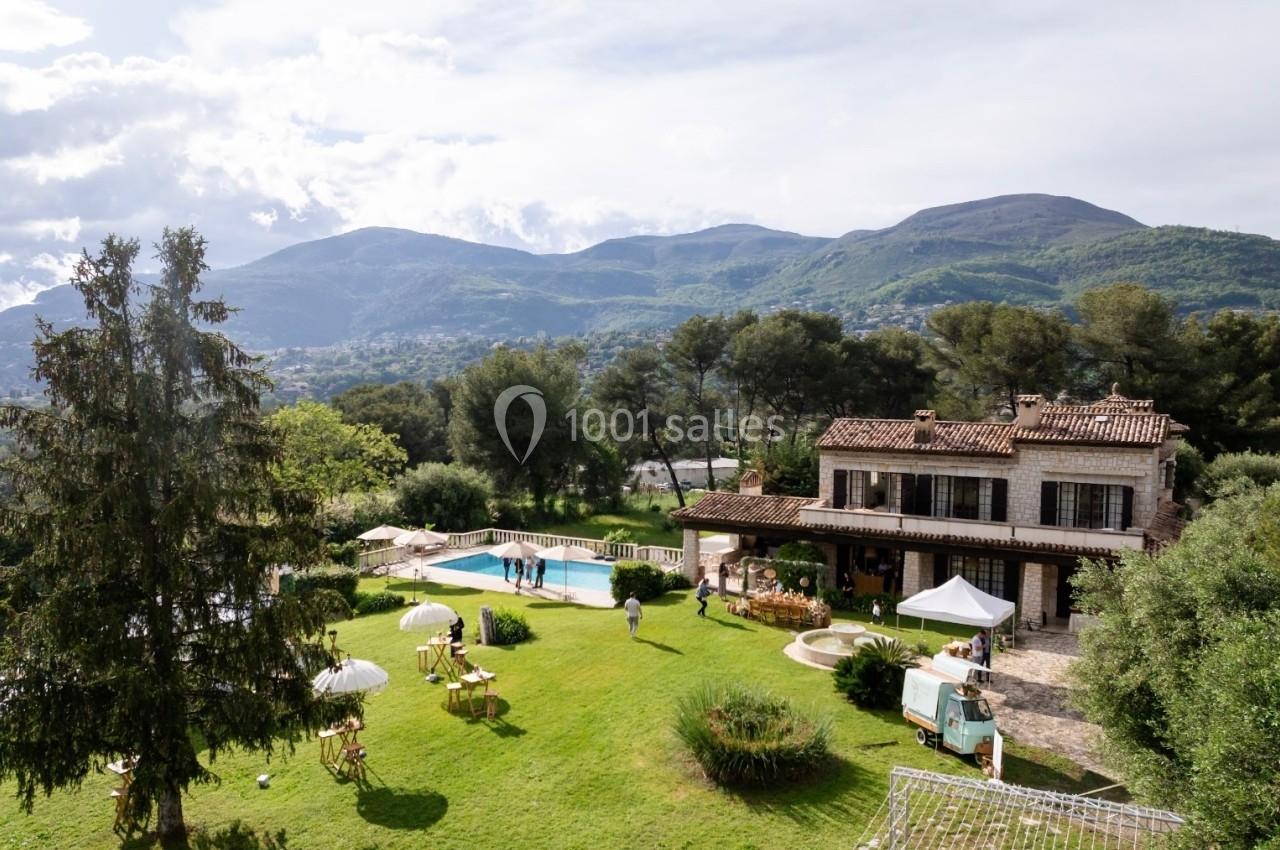 Vue d'une villa avec piscine entourée d'un grand jardin, des collines verdoyantes en arrière-plan.