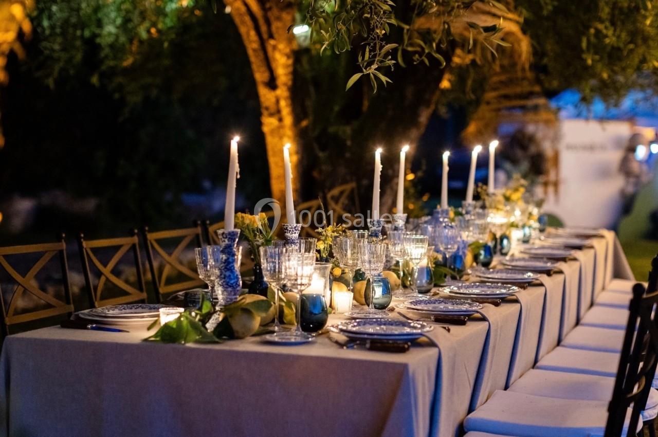 Grande table dressée en extérieur, éclairée par des bougies, avec vaisselle élégante et décorations végétales.