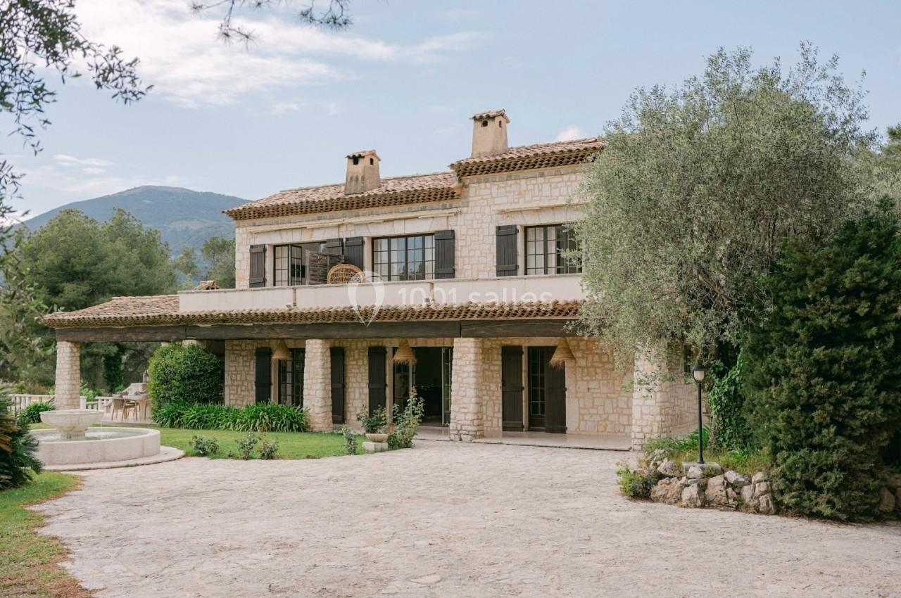 Grande maison en pierre avec terrasse, entourée de végétation, située au pied d'une colline sous un ciel dégagé.