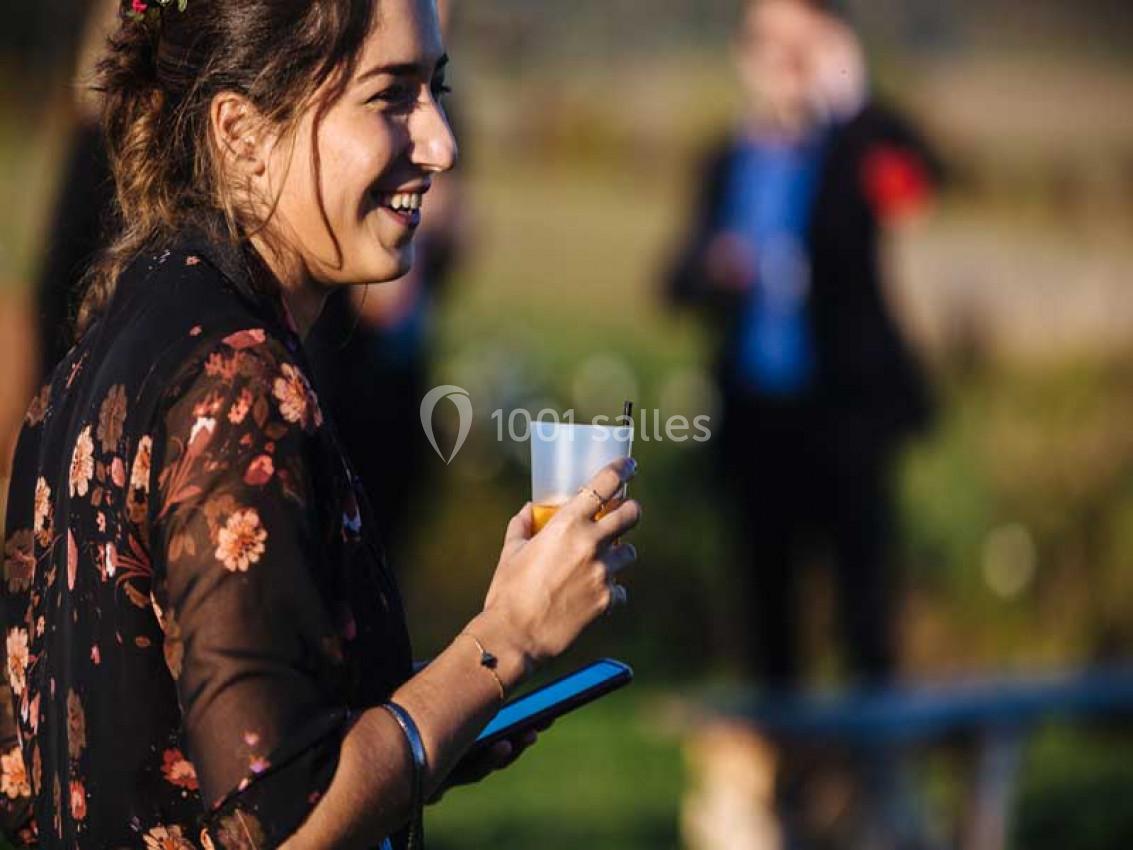 Traiteur Paris (Paris) - Profumo Traiteur Italien #20 Femme souriante tenant un verre et un téléphone dans un espace extérieur ensoleillé avec des personnes en arrière-plan flou.