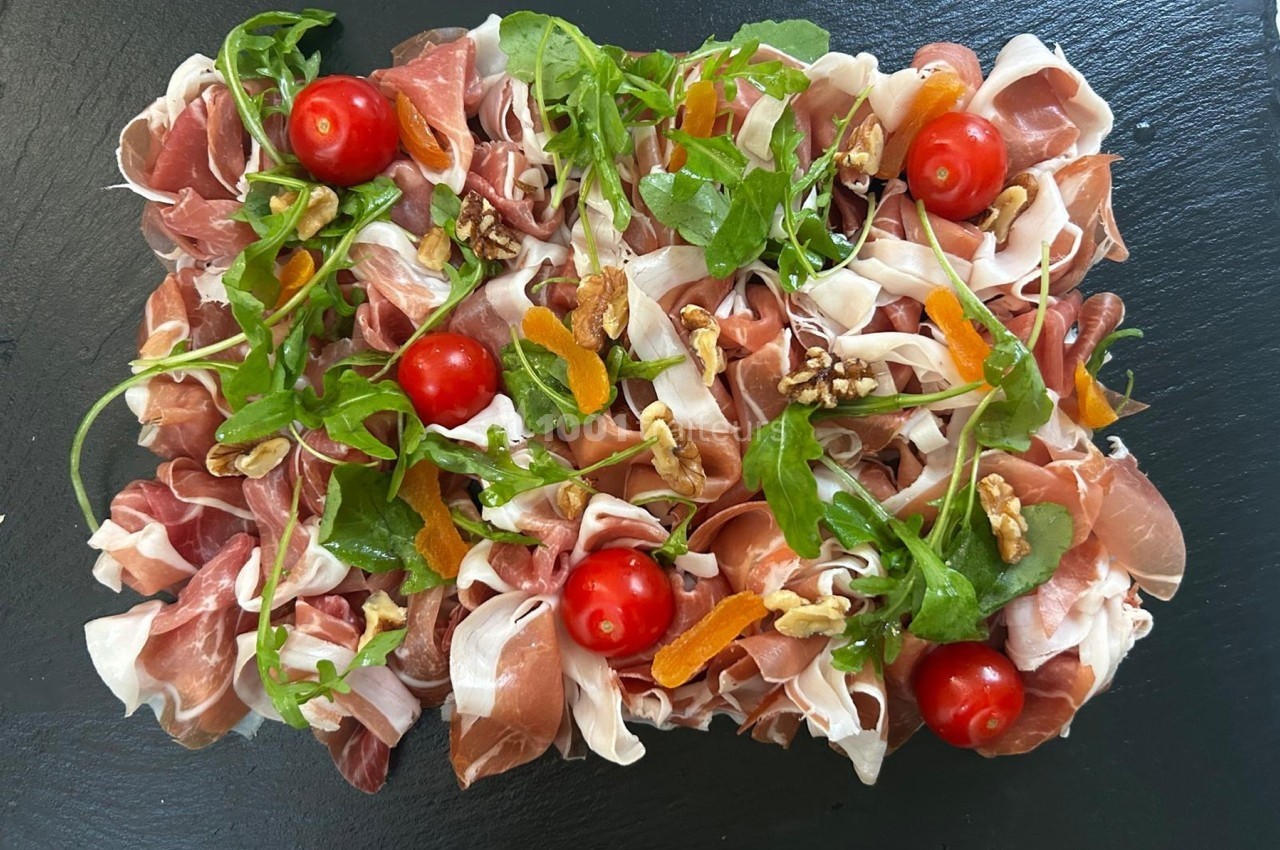 Plateau de charcuterie avec jambon cru, roquette, tomates cerises, noix et morceaux d'abricots secs sur ardoise noire.