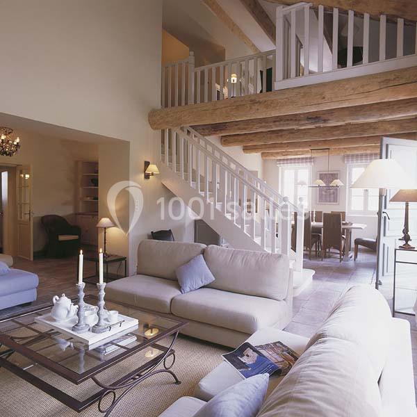 Location salle Sorgues (Vaucluse) - Château La Tour Vaucros #9 Salon lumineux avec poutres apparentes, canapé beige, table basse en verre et escalier menant à une mezzanine.
