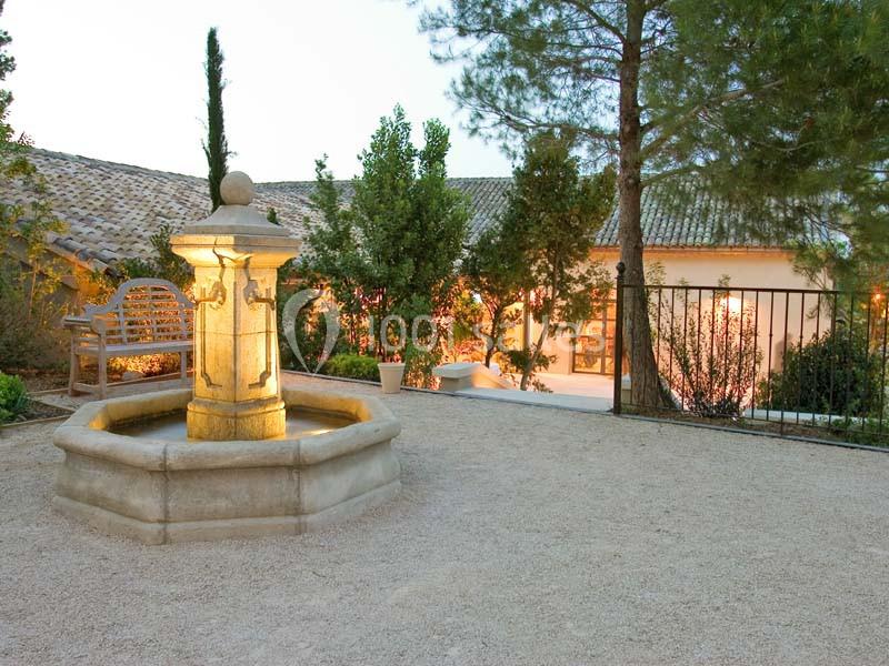 Location salle Sorgues (Vaucluse) - Château La Tour Vaucros #18 Fontaine en pierre éclairée dans une cour gravillonnée entourée de végétation et d'une maison en arrière-plan.