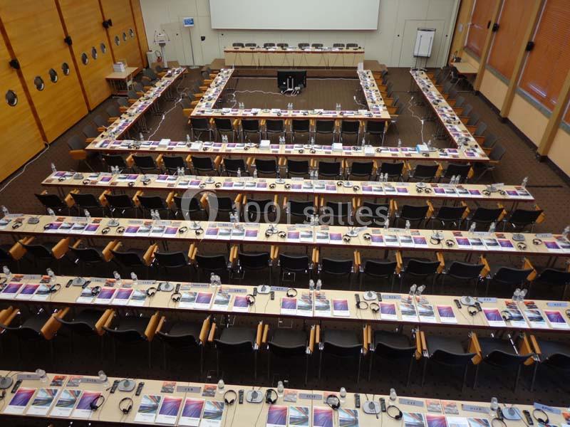 Salle de conférence vide avec des rangées de tables équipées de casques, micros et documents colorés.