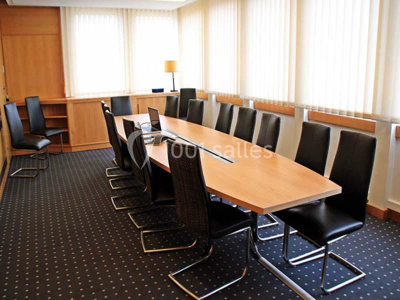 Salle de réunion avec une table ovale en bois, des chaises noires et des fenêtres avec des stores blancs.