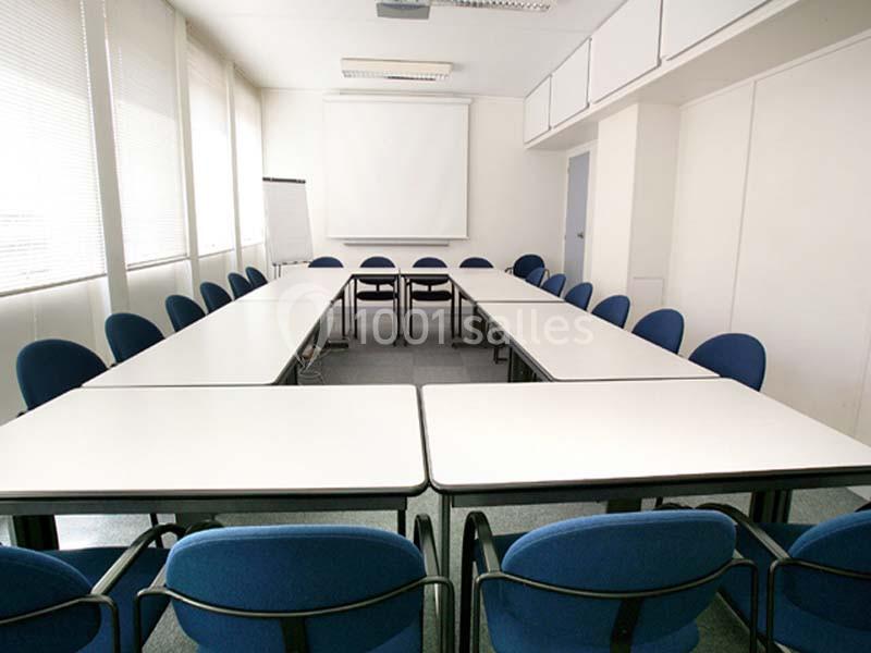 Salle de réunion lumineuse avec une grande table en U, des chaises bleues et un écran de projection.