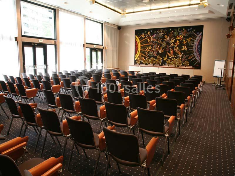 Salle de conférence vide avec rangées de chaises noires, tableau mural coloré et grandes fenêtres laissant entrer la lumière.