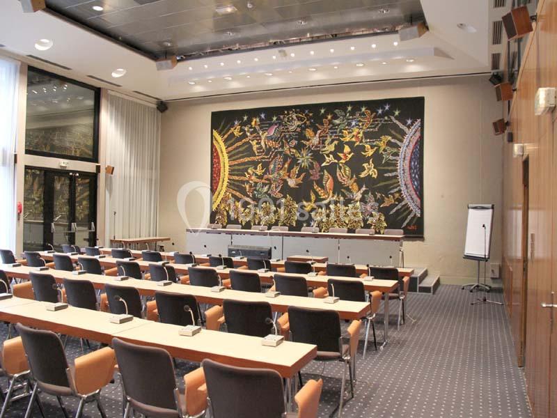 Salle de conférence avec rangées de chaises, tables équipées de micros, tableau blanc et grande tapisserie murale colorée.