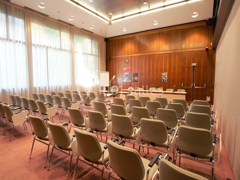 Salle de conférence vide avec des rangées de chaises beiges, un bureau en bois et un tableau blanc devant.