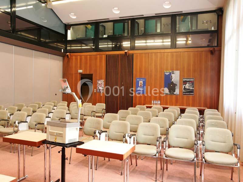 Salle de conférence vide avec des rangées de chaises beiges, un pupitre et des affiches sur les murs en bois.