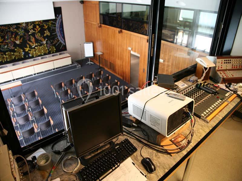 Vue depuis une régie technique sur une salle de conférence vide avec des rangées de sièges et un tableau blanc.