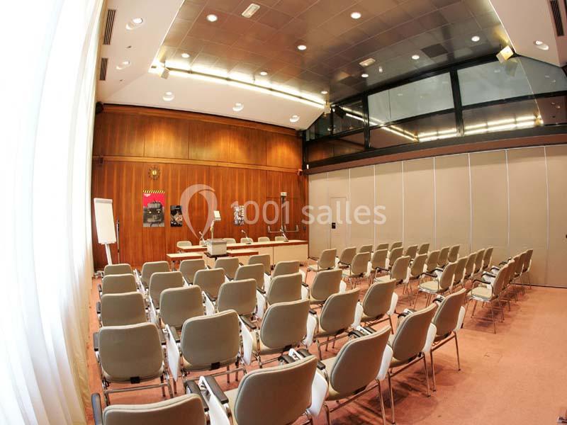 Salle de conférence avec rangées de chaises beiges, table de présentation et murs en bois.