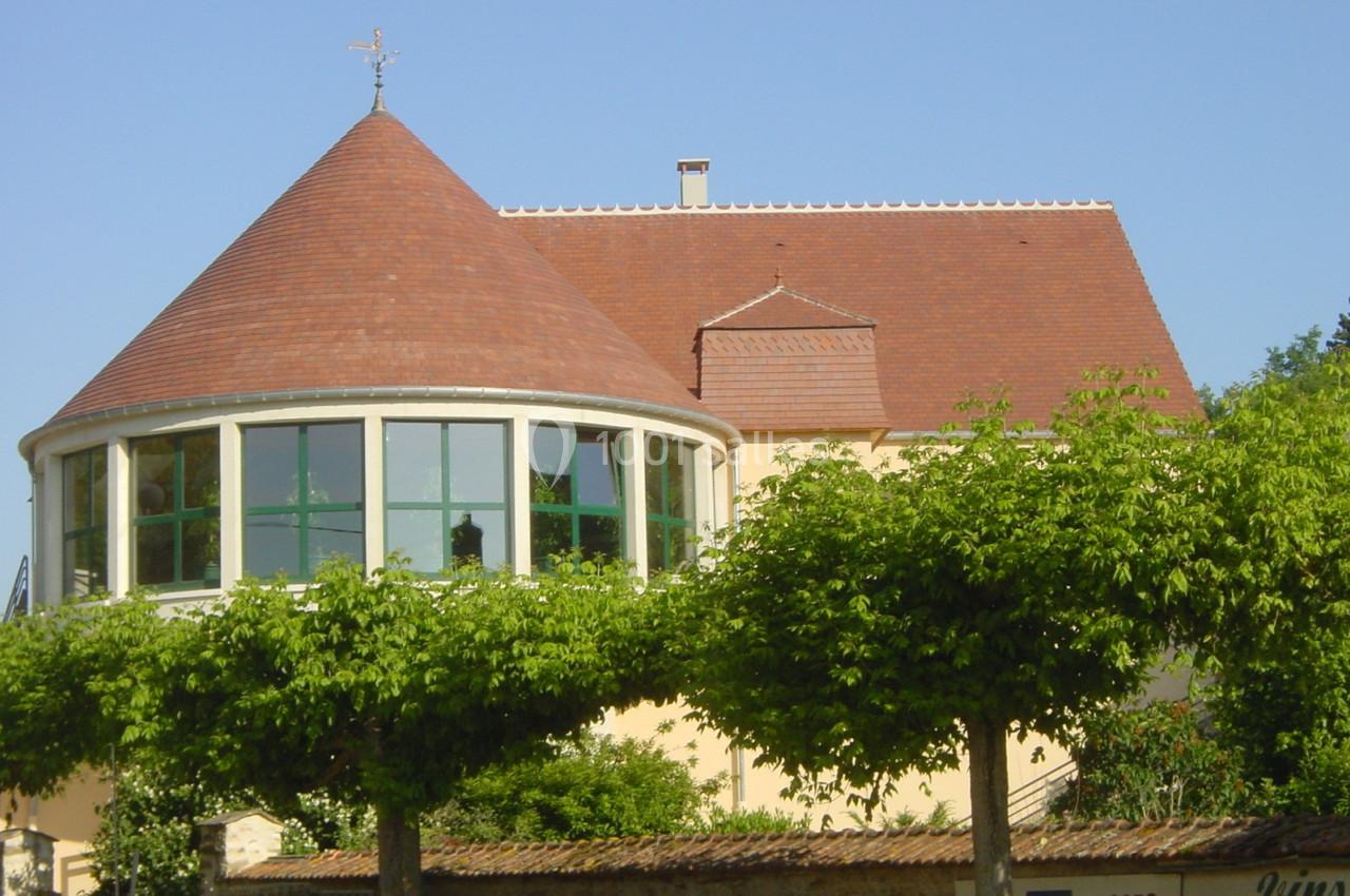 Bâtiment avec toit en tuiles rouges, grande baie vitrée arrondie et arbres taillés en avant-plan.