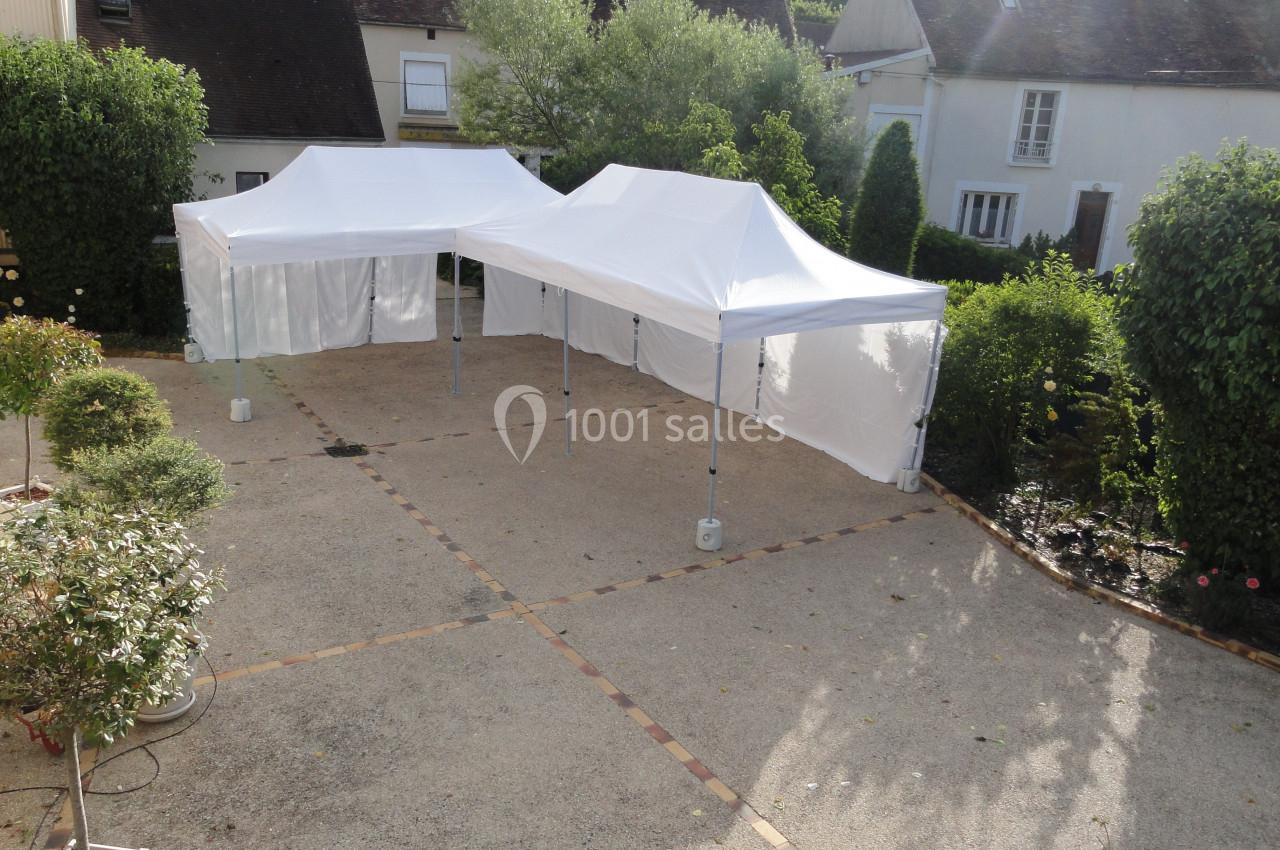 Deux tentes blanches montées sur une terrasse pavée entourée de végétation et de maisons.