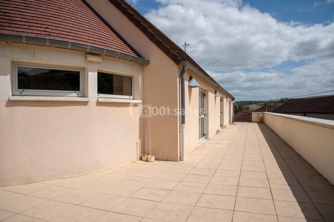 Grande terrasse en carrelage beige longeant un bâtiment aux murs clairs et toit en tuiles, sous un ciel partiellement…