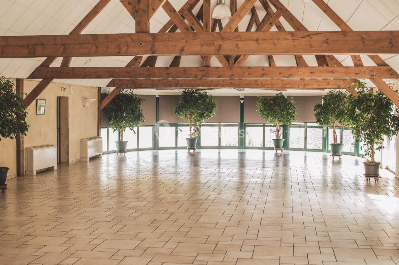 Salle spacieuse avec charpente en bois apparente, carrelage clair et plantes en pots alignées près des fenêtres.