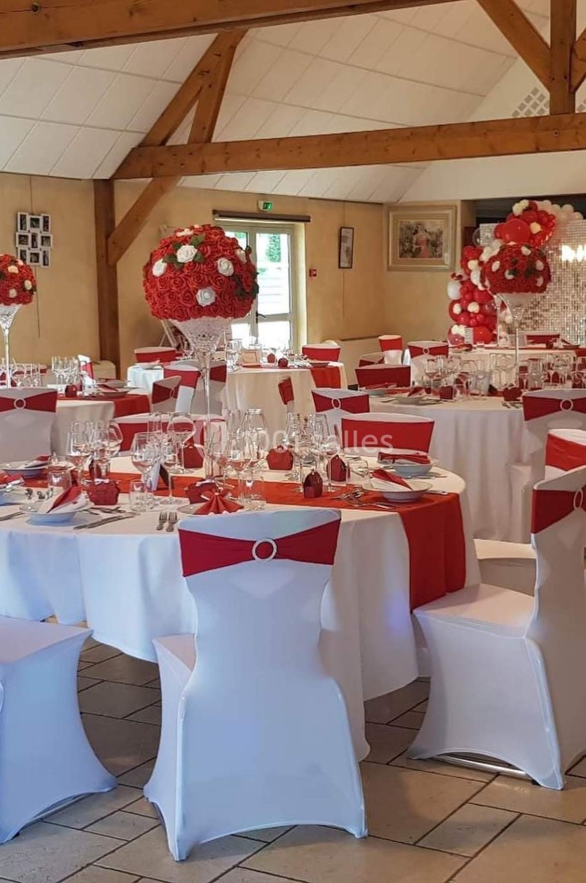 Salle de réception décorée avec des tables rondes, nappes blanches et rouges, et centres de table floraux assortis.