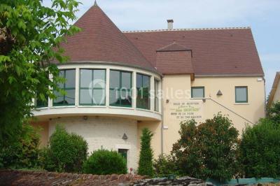 Salle spacieuse avec charpente en bois apparente, carrelage clair et plantes en pots alignées près des fenêtres.