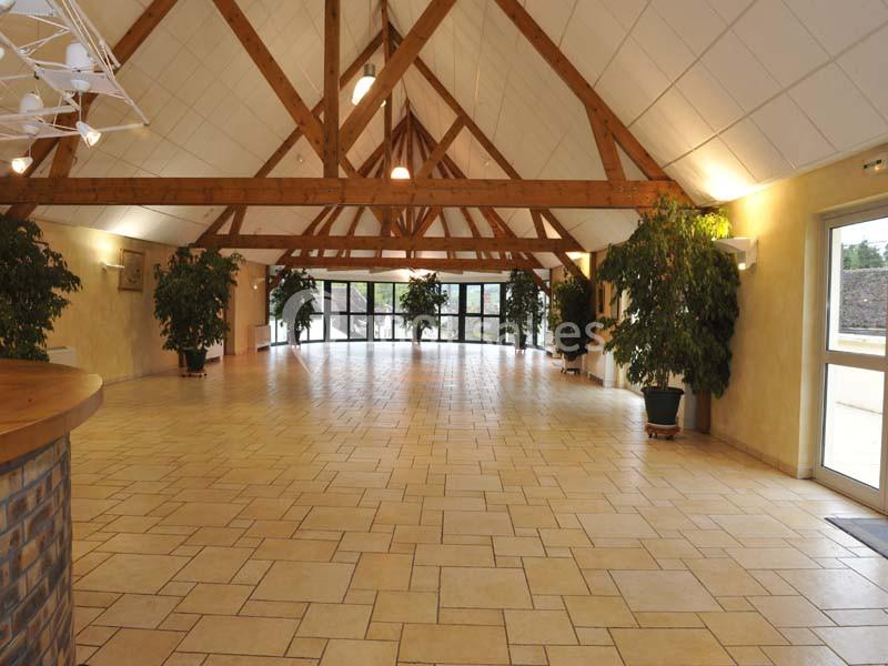 Salle spacieuse avec charpente apparente en bois, carrelage clair, grandes fenêtres et plantes en pot.