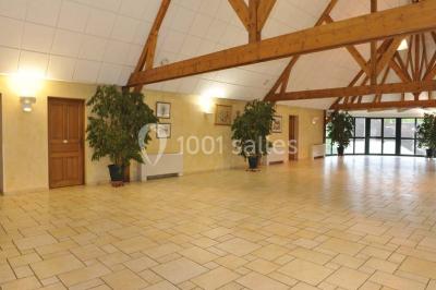Salle spacieuse avec charpente en bois apparente, carrelage clair et plantes en pots alignées près des fenêtres.