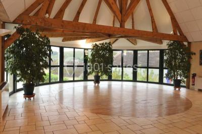 Salle spacieuse avec charpente en bois apparente, carrelage clair et plantes en pots alignées près des fenêtres.