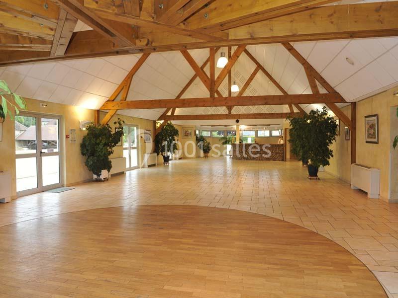 Salle spacieuse avec charpente en bois apparente, sol en parquet et carrelage, entourée de grandes fenêtres et de plantes.