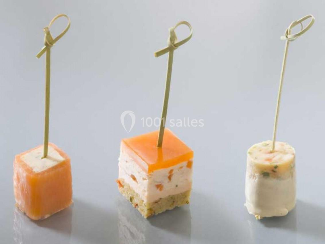 Trois amuse-bouches sur piques en bois, composés de saumon, mousse et autres ingrédients, disposés sur une surface claire.