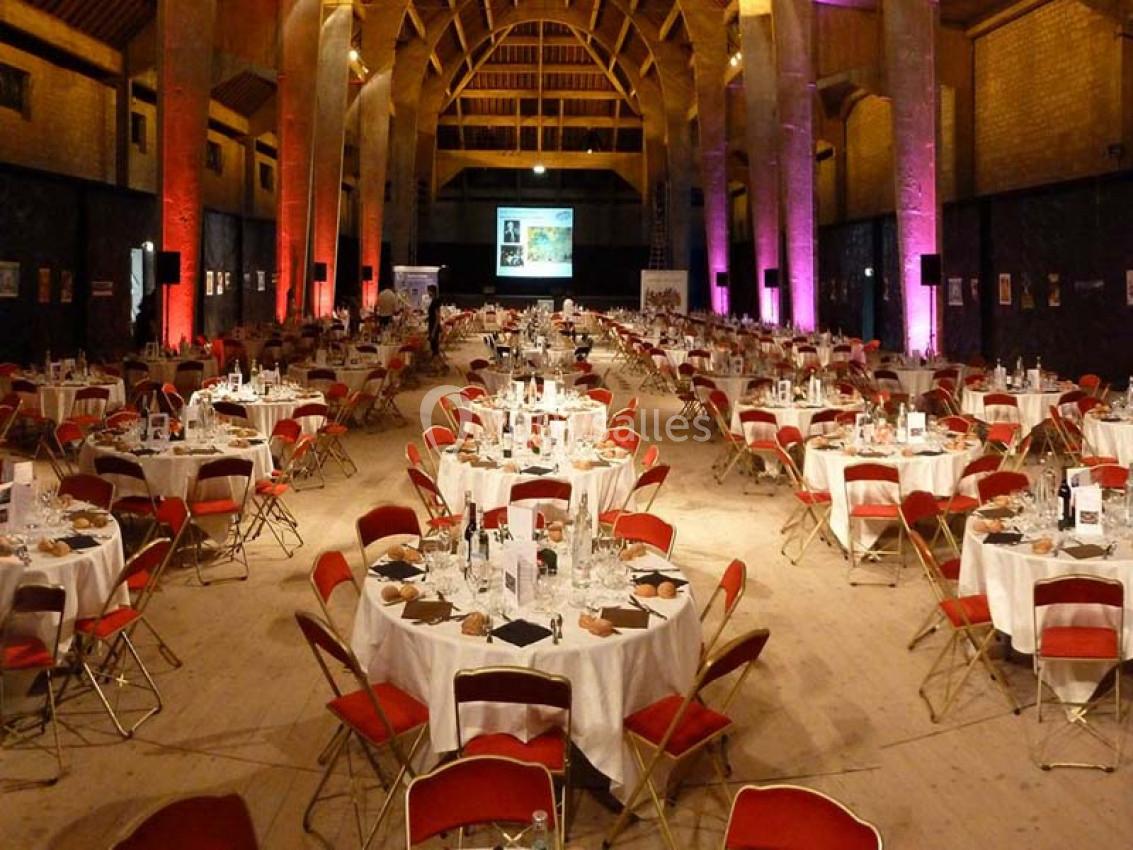 Salle de réception avec de longues rangées de tables rondes dressées, entourées de chaises rouges, sous une charpente en…