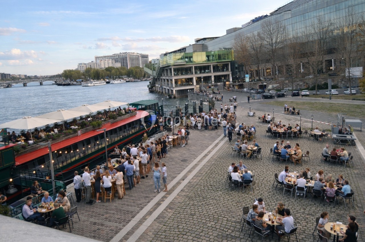 Terrasse animée avec des tables en plein air près de la Seine, des passants et une péniche aménagée en restaurant.