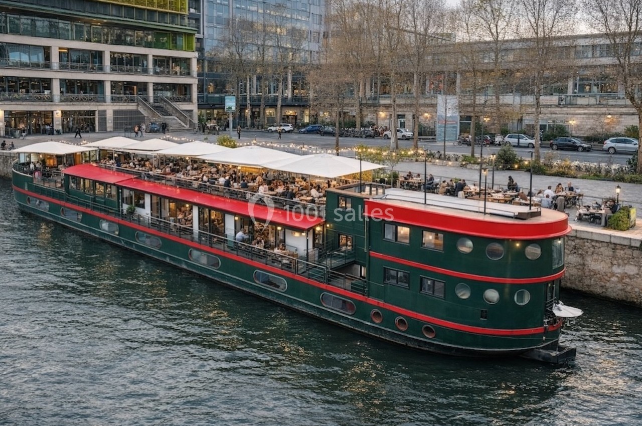 Bateau aménagé en restaurant avec terrasse couverte, amarré le long d'un quai bordé d'arbres et de bâtiments modernes.