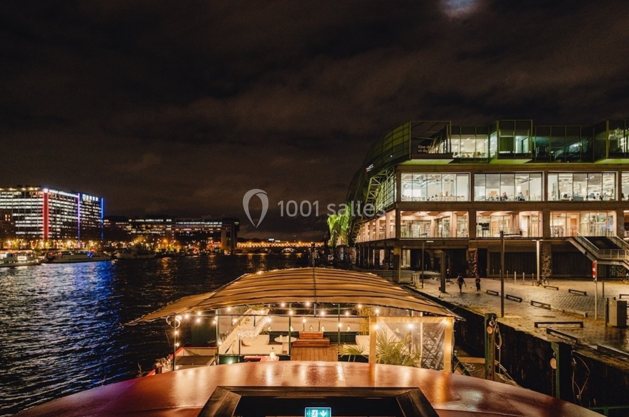 Vue nocturne d'un quai urbain avec un bâtiment moderne éclairé et un bateau amarré au premier plan.