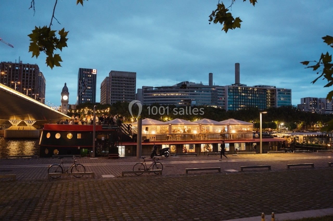 Vue nocturne d'une péniche éclairée amarrée près d'une rive, avec des bâtiments modernes en arrière-plan.