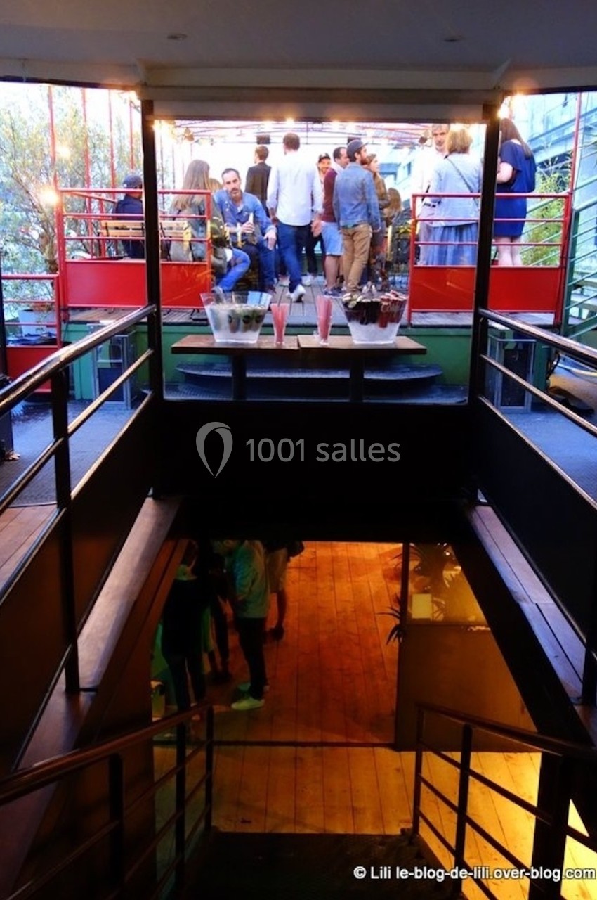 Vue d'un escalier menant à une terrasse animée avec des personnes discutant autour de tables et de plantes.