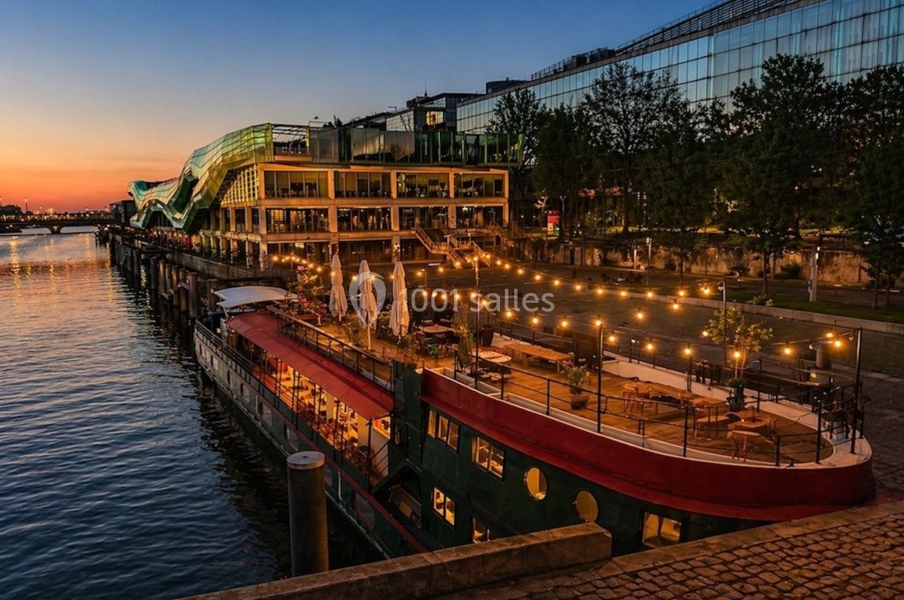 Bateau-restaurant éclairé amarré le long d'un quai, avec bâtiments modernes et ciel au crépuscule en arrière-plan.