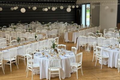 Salle de réception décorée avec des tables rondes et rectangulaires, nappes blanches, chaises blanches et guirlandes…