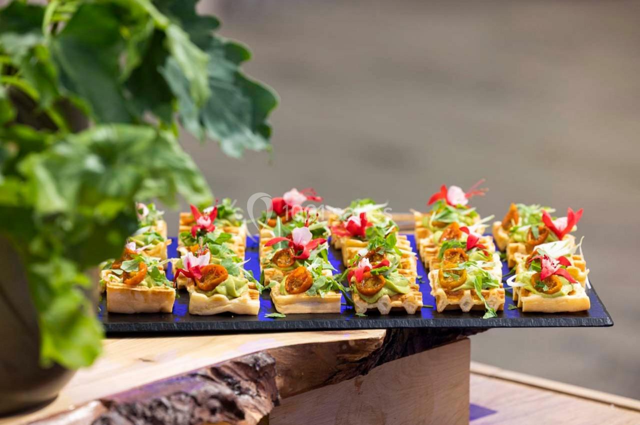 Plateau de mini-gaufres garnies de légumes, herbes fraîches et fleurs comestibles, posé sur une table en bois.