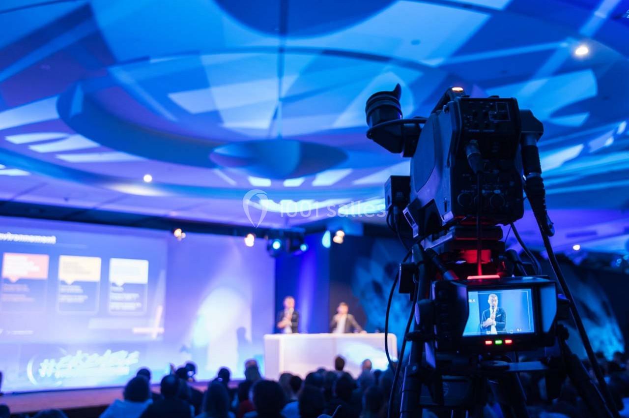 Caméra filmant une conférence sur scène, avec deux intervenants et un public dans une salle éclairée.