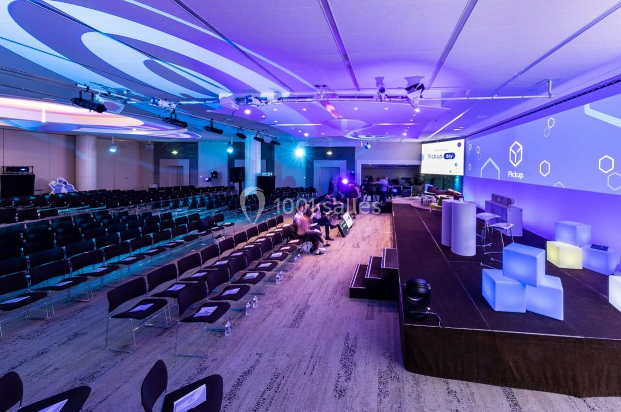 Salle de conférence moderne avec des sièges alignés, une scène équipée et des écrans affichant des visuels lumineux.