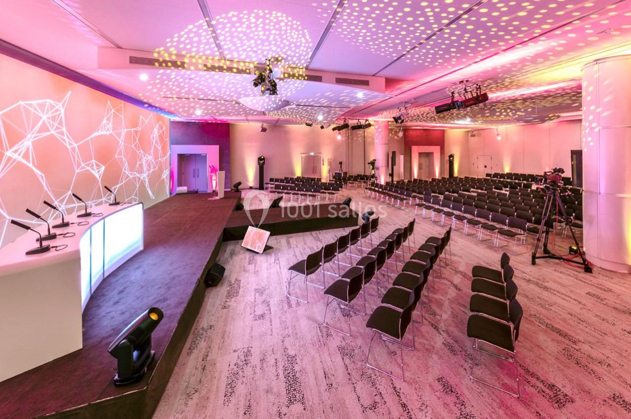Salle de conférence moderne avec rangées de chaises, scène équipée de micros et éclairage coloré.