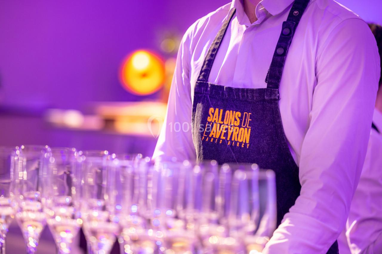 Serveur en tablier portant l'inscription ’Salons de l'Aveyron’ devant une rangée de verres à champagne.