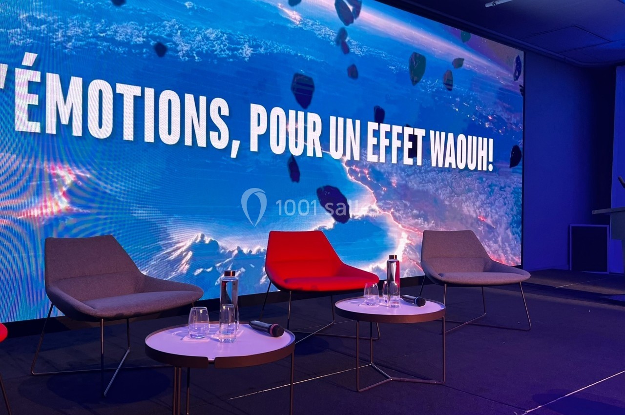 Quatre fauteuils et trois tables basses sur une scène, devant un écran affichant un message et un fond coloré.