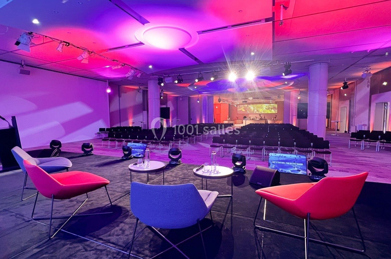 Salle de conférence moderne avec chaises colorées sur scène, éclairage violet et rangées de sièges vides en arrière-plan.