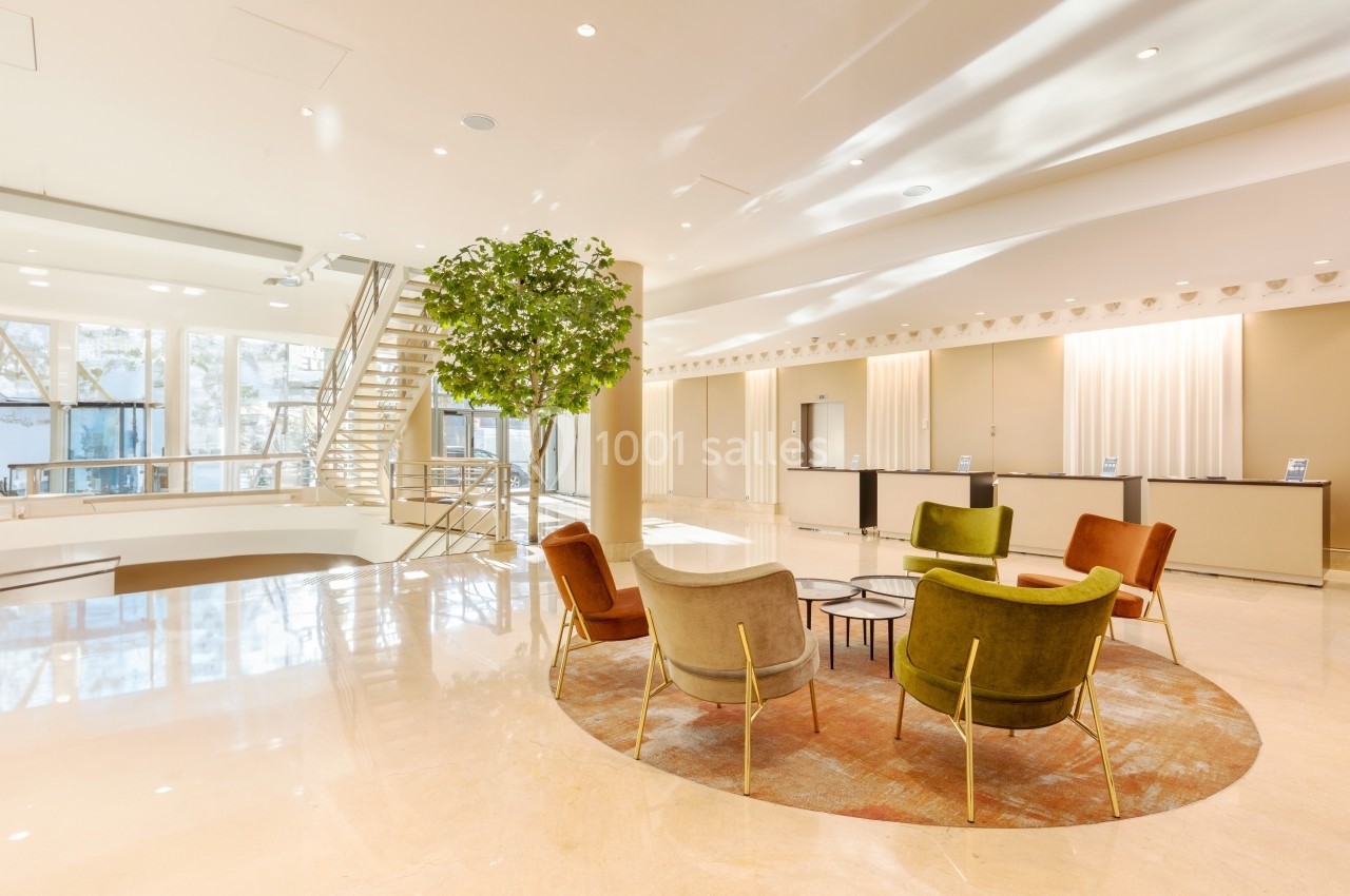 Hall lumineux avec des fauteuils colorés autour d'une table basse, un tapis rond et un escalier en arrière-plan.