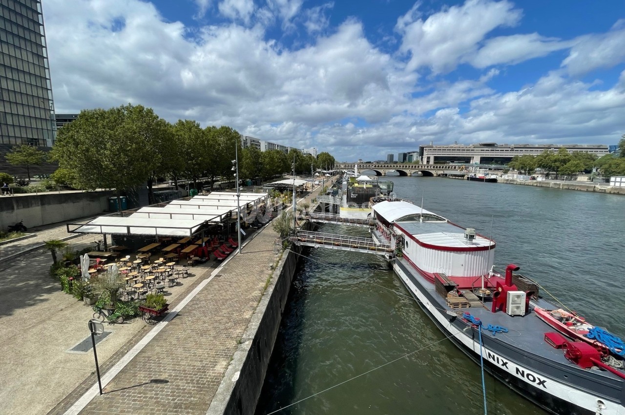 Terrasse fluviale avec voiles blanches, chaises en bois, bateau rouge et blanc amarré au quai fluvial parisien.