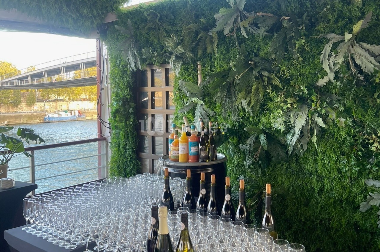 Espace ouvert au bord de l'eau avec murs de végétation, verres vides et bouteilles de vin alignées.