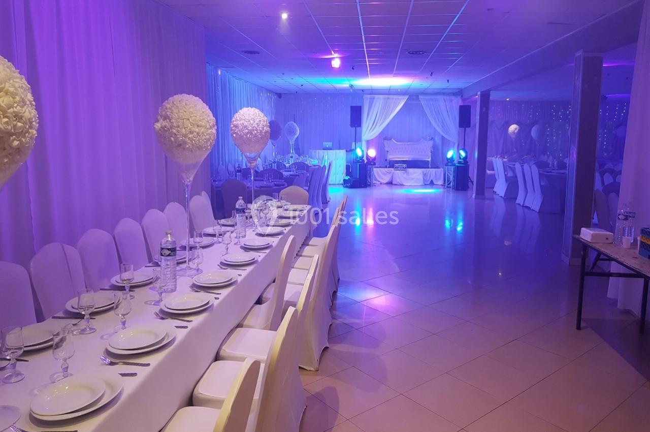 Salle de réception décorée avec des tables dressées, chaises blanches et éclairage bleu-violet.